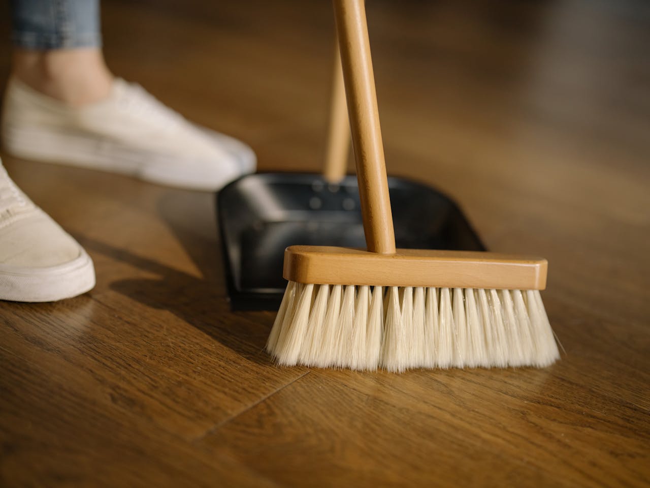 Close-up of a broom and dustpan with white sneakers indoors, representing housekeeping and cleanliness.